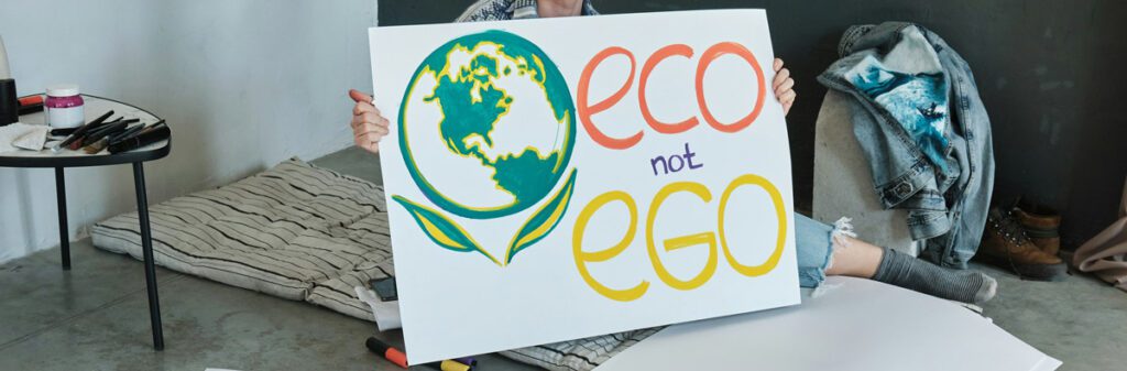 A person holds a sign featuring a drawing of Earth and the words "eco not ego" in colorful letters. The individual, an advocate for climate change awareness, is seated on the floor beside art supplies with a casual outfit draped over a nearby chair. The setting suggests a cozy, creative space, perfectly capturing the message of "Header_Excerpt_What-does-climate-hypocrite-mean".