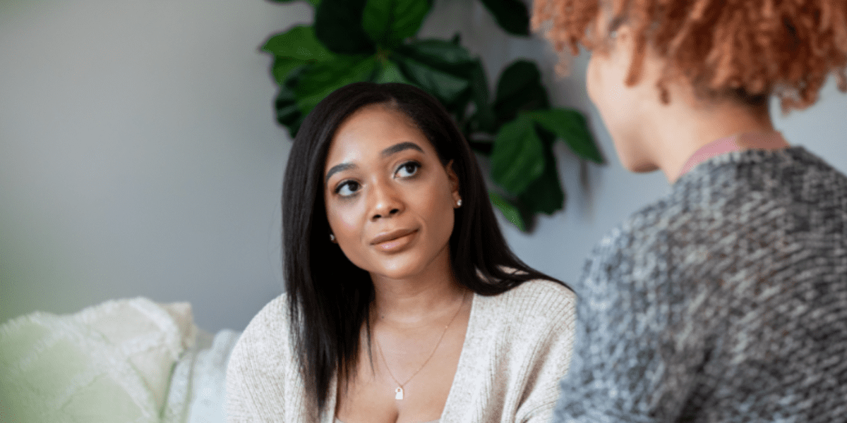 Two young women sit on a couch having a conversation.