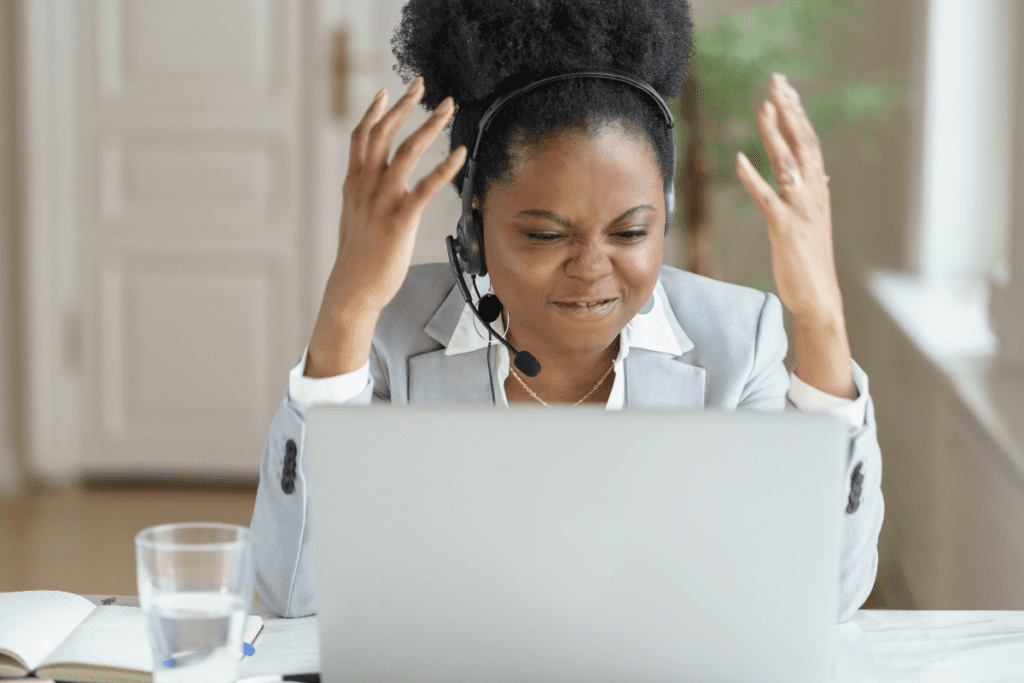 A woman at a computer wearing a headset raises her arms in frustration.