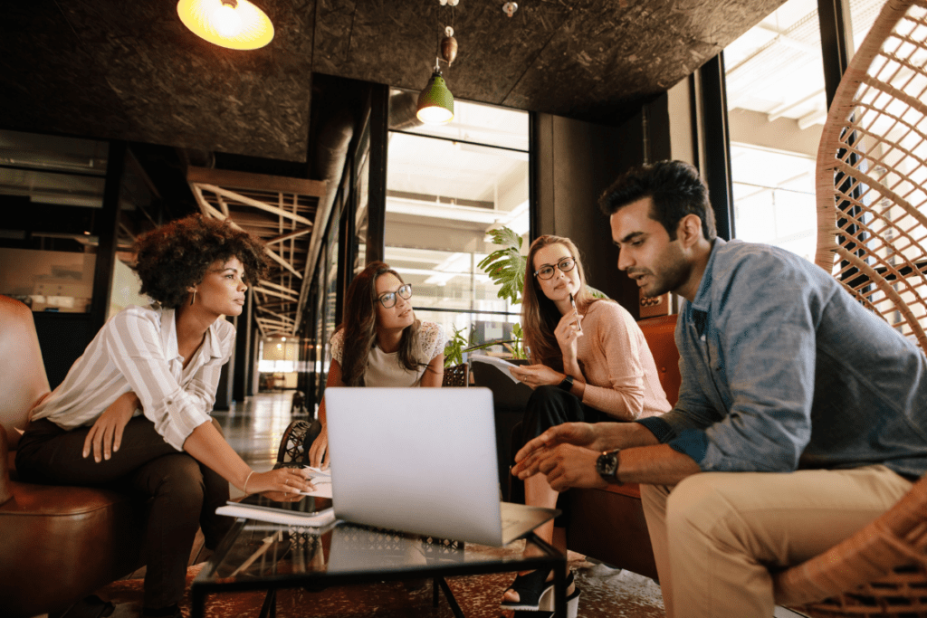 Three women and one man have a conversation around a coffee table with a laptop open.