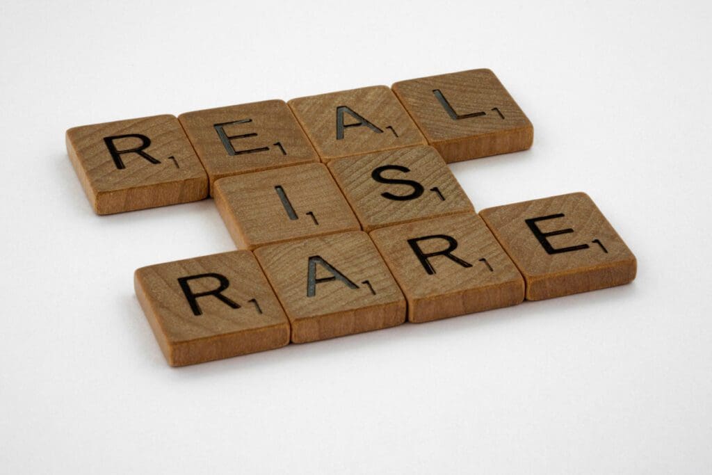 Wooden Scrabble tiles from the brett-jordan-zVkL5L7eGrw-unsplash collection are arranged on a white background to spell out "REAL IS RARE." The word "REAL" intersects with "IS" and "RARE" in a crossword-like layout. Each tile has a single point value of 1, subtly reminding us that confronting environmental hypocrisy is crucial in addressing climate change.