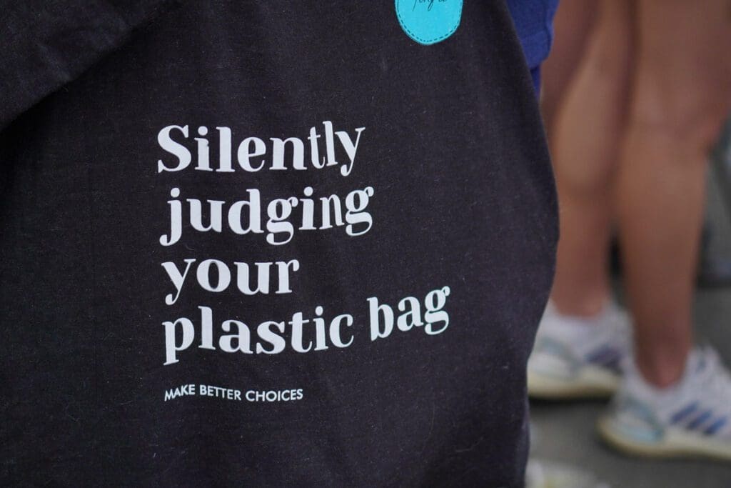 Close-up of a person wearing a black shirt featuring white text that reads, "Silently judging your plastic bag" and "Make better choices." In the blurred background, people in casual attire are visible, including legs and feet in athletic shoes. It's a nod to environmental accountability in everyday choices. 