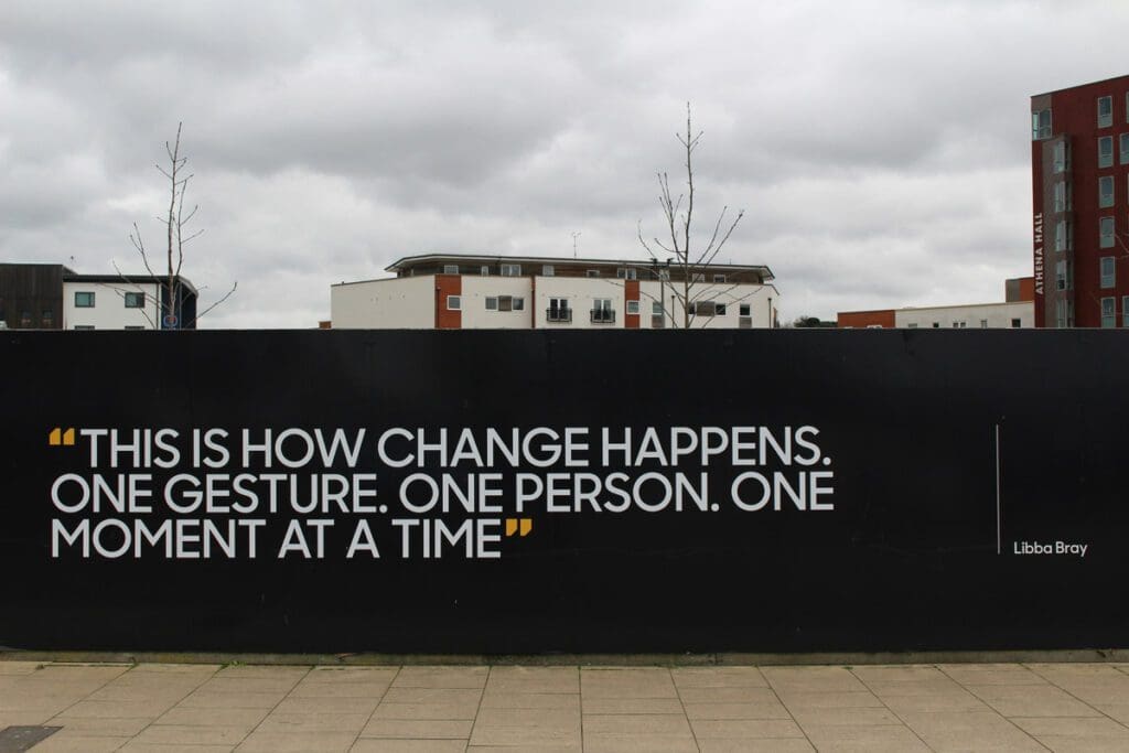 A black wall, part of the "david-walker-bP0q3tTTHS4-unsplash" piece, displays the quote "This is how change happens. One gesture. One person. One moment at a time." by Libba Bray in white letters against a backdrop featuring a cloudy sky and modern buildings, subtly reminding us of the urgency of addressing climate change amidst growing hypocrisy.