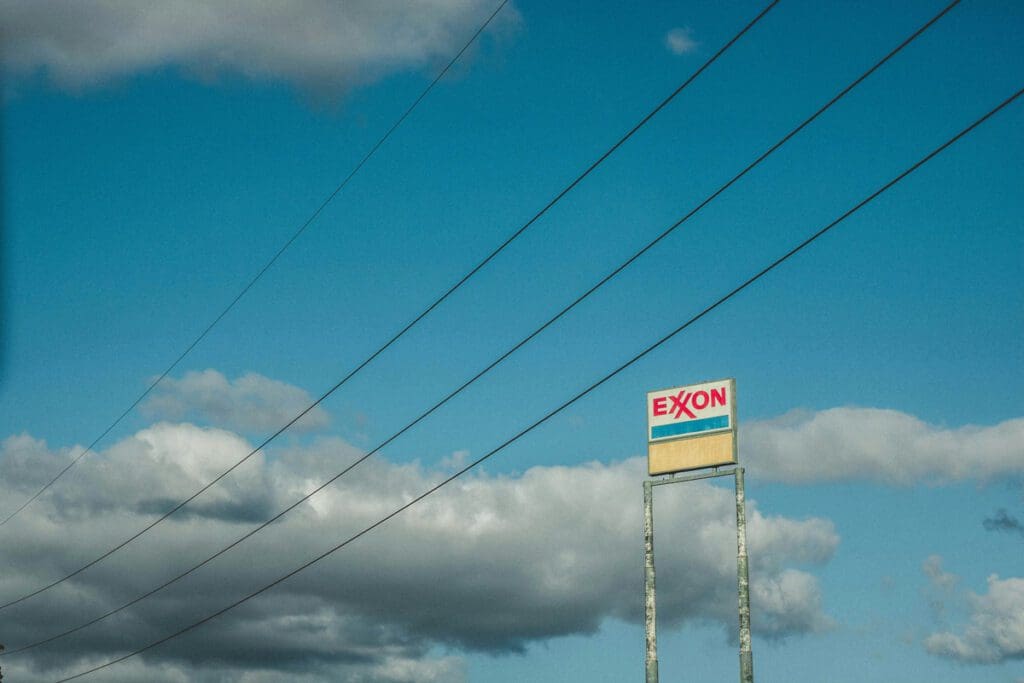 A photograph titled "justin-6bsVqTJna_o-unsplash" captures an Exxon gas station sign standing tall against a backdrop of blue sky and fluffy clouds, with several power lines cutting diagonally across the image, subtly reminding us of the ongoing challenge of climate change.