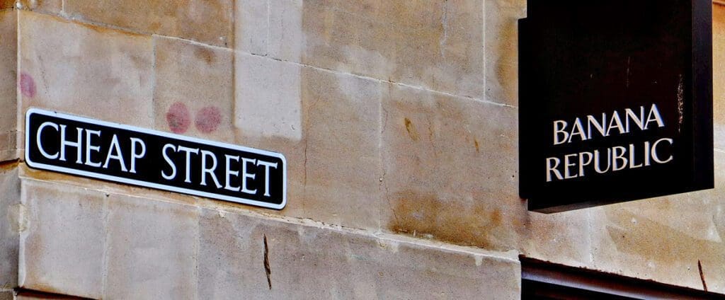 A street sign reading "CHEAP STREET" is mounted on a beige stone wall. Adjacent to it, there is a black store sign for "BANANA REPUBLIC," also attached to the wall, creating an ironic juxtaposition that could make one ponder the subtle hypocrisies of urban branding, as captured in kerry-rawlinson-TNl4aR3kidM-unsplash.