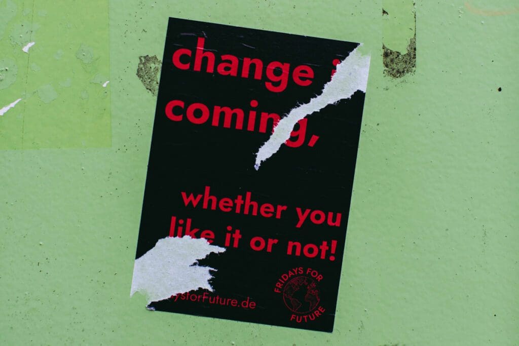 A partially torn poster, identified as "markus-spiske--iOE0qBkioc-unsplash," featuring bold red text on a black background hangs on a light green wall. It reads: "change is coming, whether you like it or not! Fridays For Future." Despite the jagged tears that mar the text and give it a defiant tone toward climate hypocrites, the website "fridaysforfuture.de" remains visible below.