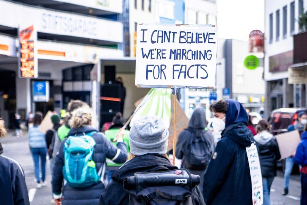 A group of protesters march down a city street. One person holds a sign that reads, "I can't believe we're marching for facts." They wear backpacks, jackets, and some have face masks on. Business buildings and signs are visible in the background as they rally against climate change hypocrisy, similar to the scene captured in mika-baumeister-wcv_MlJtRYA-unsplash.