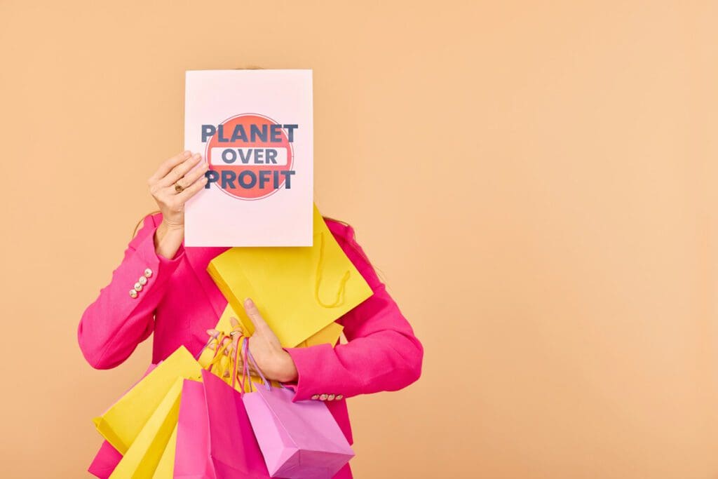 A person in a bright pink blazer, holding multiple colorful shopping bags and an A4 sign with the text "PLANET OVER PROFIT" in front of their face, is captured in natalia-blauth-n7GUZ_nV_LM-unsplash, emphasizing the irony of climate hypocrisy against a solid beige background.