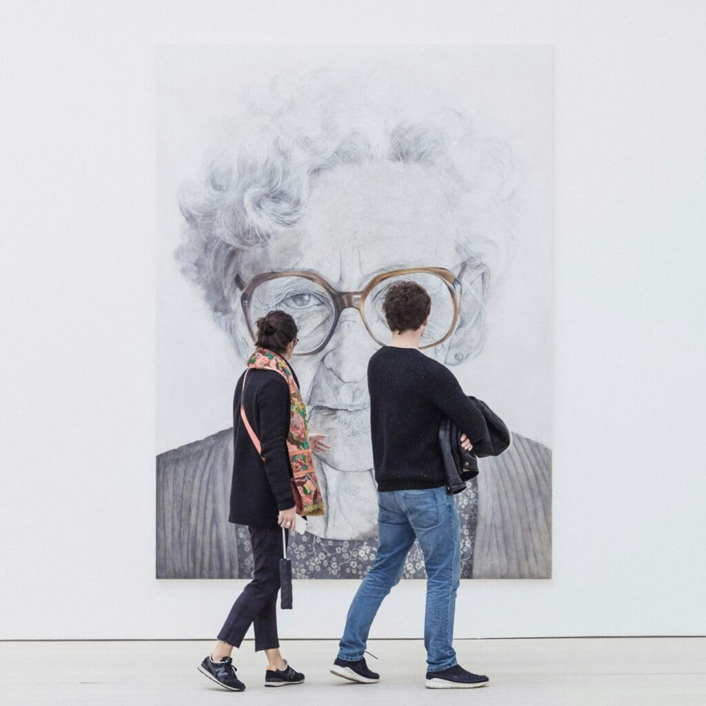Two individuals are walking past "ryan-stefan-5K98ScREEUY-unsplash," a large, gray-toned portrait of an elderly person with curly hair and large glasses. The portrait is displayed on a white wall. One person is wearing a black top and colorful scarf, while the other in a black sweater, quietly discussing climate change.