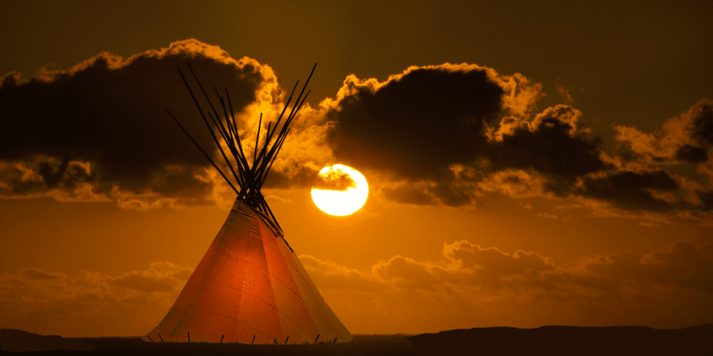 Silhouette of a teepee with a vibrant sunset and clouds in the background.