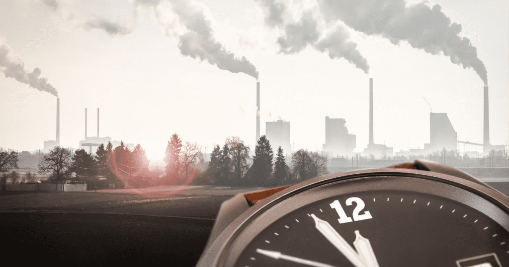 A clock foregrounds a hazy skyline with large smokestacks emitting smoke, reminiscent of an auto draft from yesteryears' industries.