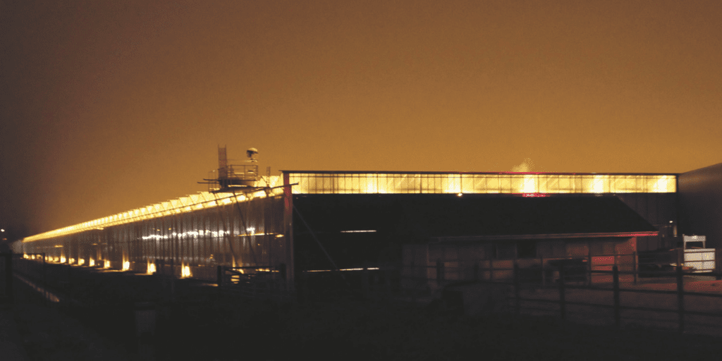 The greenhouse, a simple shelter with warm, yellow lights, glows against a hazy orange sky at night, ideal for winter farming to protect crops.