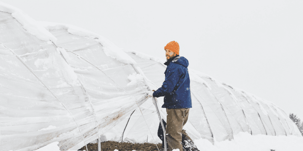 A person in a blue jacket and orange hat adjusts a snow-covered greenhouse, making effective use of a crop shelter to safeguard crops in the snowy landscape. Winter farming at its finest.