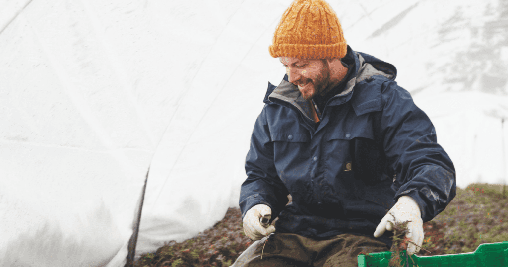 A person in a winter coat and beanie smiles while harvesting crops in a greenhouse, beautifully illustrating the blend of nature's warmth with the efficiency.