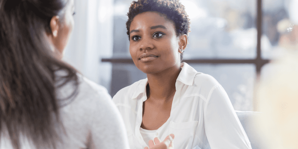 Two women engage in a serious discussion: one with short curly hair in a white shirt listens attentively as the other, with long dark hair, speaks passionately.