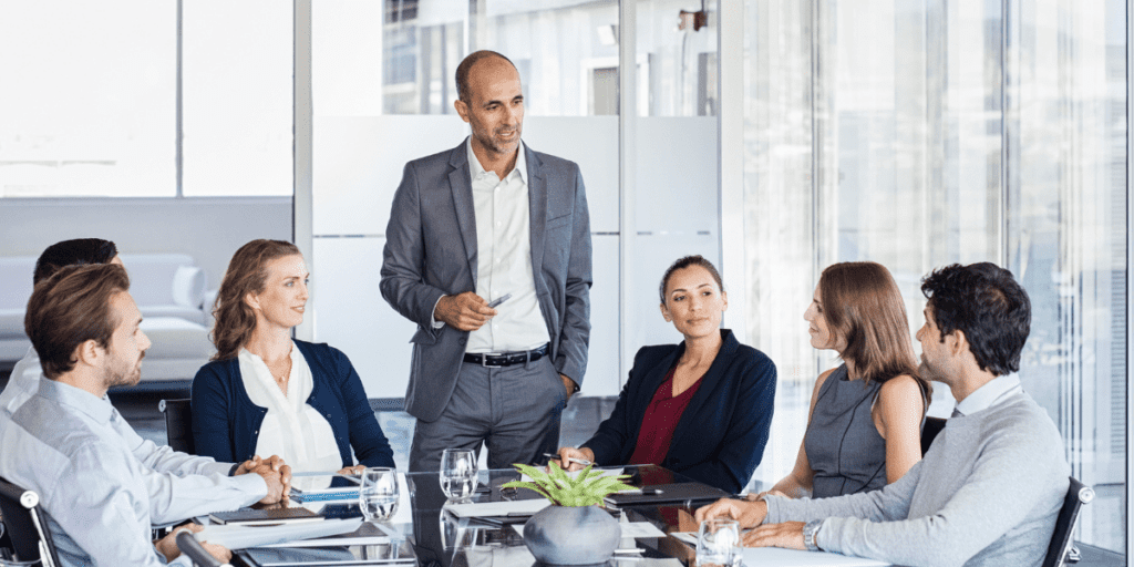 During a meeting in a modern office setting, a man in a gray suit stands and speaks to six seated colleagues, addressing strategies for resolving conflict efficiently.