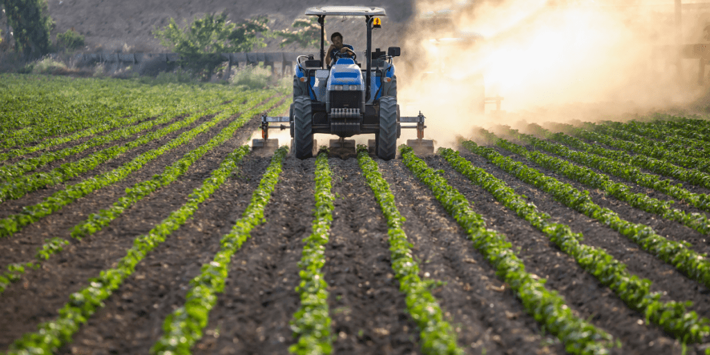 A tractor plows between rows of crops on a sunny day, with dust rising behind it, showcasing resilient agriculture techniques designed to tackle global challenges.
