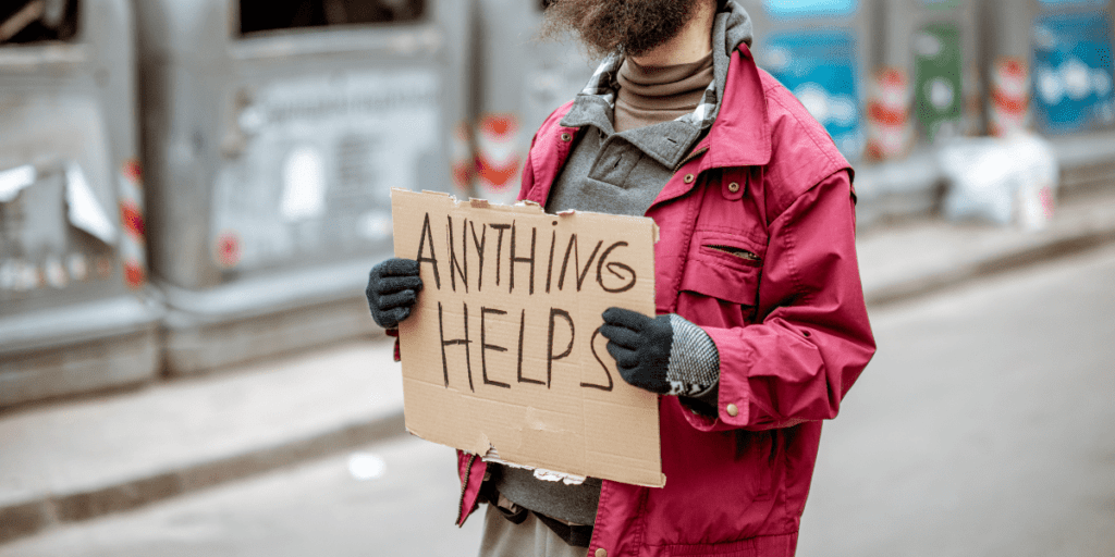 A person stands on a city street, holding a cardboard sign that reads "Anything Helps," silently bearing the invisible chains of poverty.