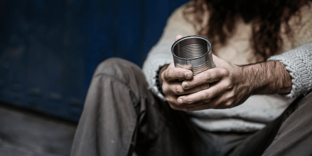 A person sits wrapped in a sweater and brown pants, holding an empty tin can.
