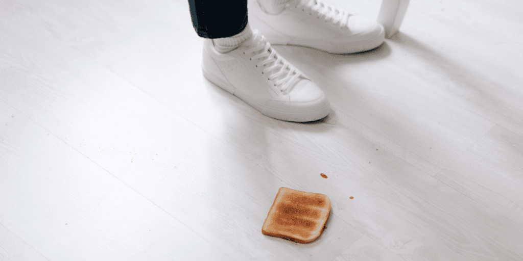 A piece of toast rests on a light wooden floor near a person's white sneakers.