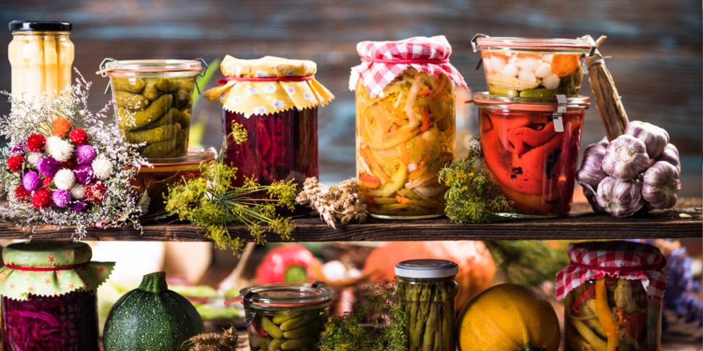 Jars of pickled vegetables and herbs, stored on a wooden shelf.