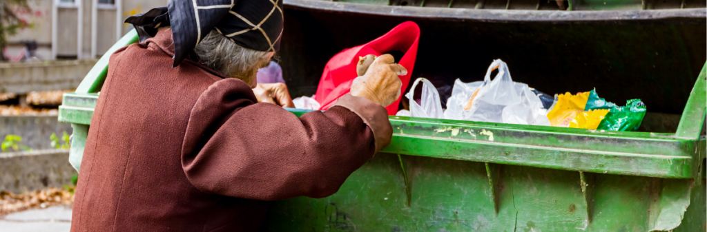 An elderly person searches through a green dumpster filled with bags.