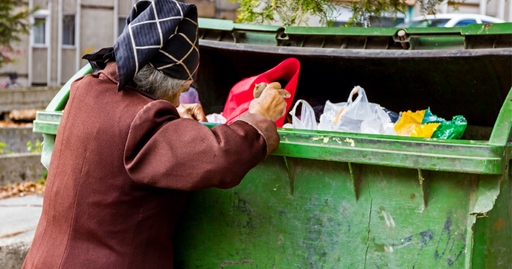 An elderly person, shackled by the invisible chains of poverty, searches through a large green dumpster outside.