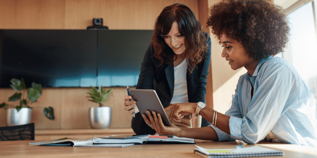 In a modern office setting, two women are collaborating over a tablet.