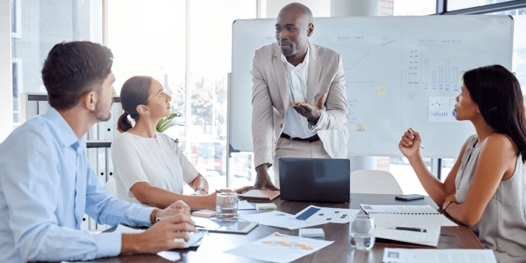 A man leads a meeting, expertly guiding three colleagues through a discussion with a whiteboard and laptops.