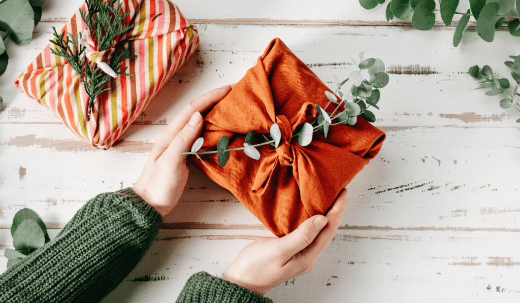 Hands holding a gift wrapped in orange fabric with eucalyptus sprigs—an eco-friendly gathering touch.