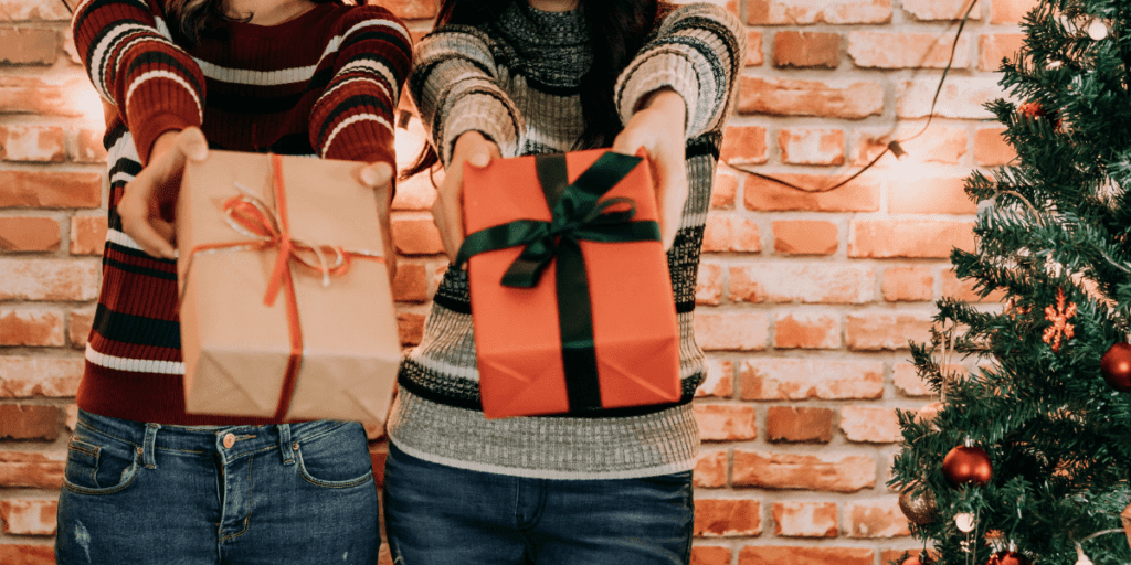 Two people holding thoughtful gifts in front of a brick wall and a decorated Christmas tree, embodying the spirit of conscious gifting.