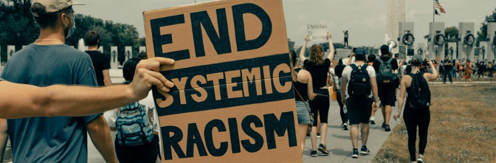 People march near a monument, engaging in racial conversations, as one holds a sign reading "End Systemic Racism.