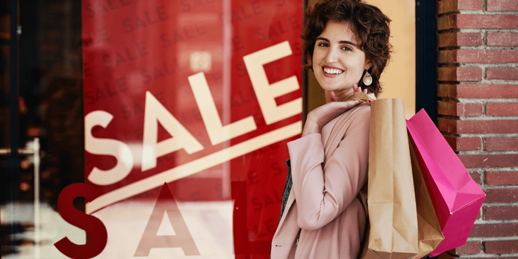 A smiling person holding shopping bags stands in front of a sale sign, having unlocked the secret to smart buying decisions this season.
