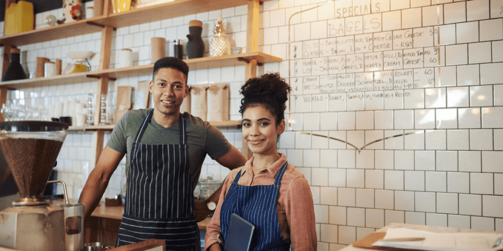 Two baristas smiling behind the counter in a cozy coffee shop, creating a warm atmosphere where patrons can support local business without spending money on anything but the perfect cup of joe.