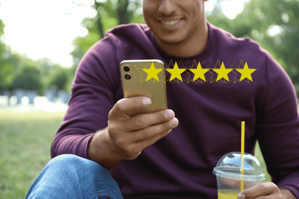 Smiling man using phone with a five-star rating overlay, supporting local businesses without spending money, all while sitting outdoors with a refreshing drink.