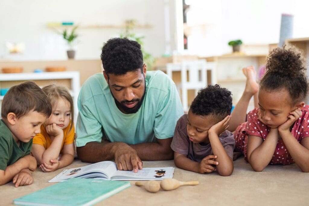 In a classroom setting, a man reads a book to four children on the floor, fostering open communication and inviting them to talk about race with curiosity and understanding.