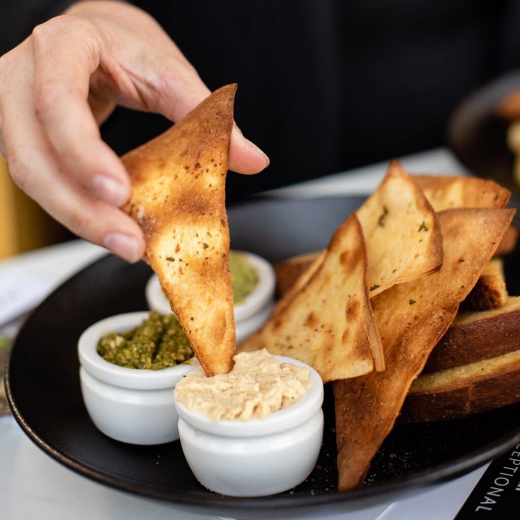 Hand dipping pita chip into hummus, with a side of crackers and other delicious spreads, perfect for Christmas gatherings.