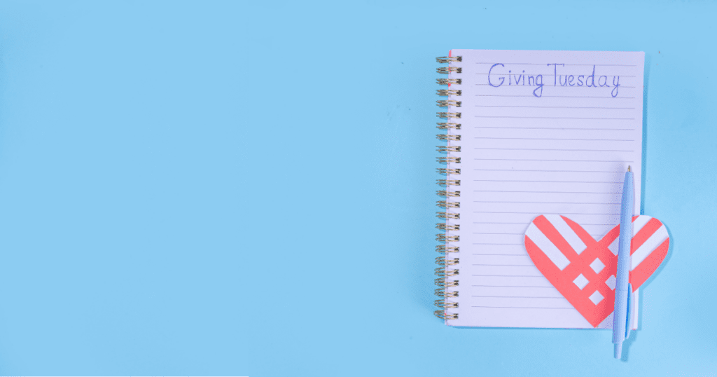 Notebook with "Giving Tuesday" written on it, a red heart, and a blue pen on a light blue background, highlighting the spirit of giving back.