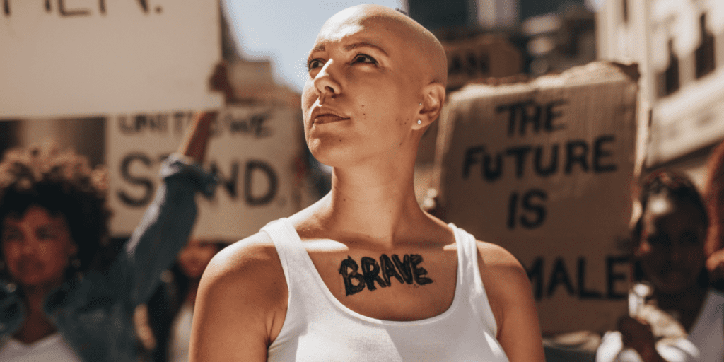 Person with "BRAVE" tattoo on chest at a protest with signs in background.