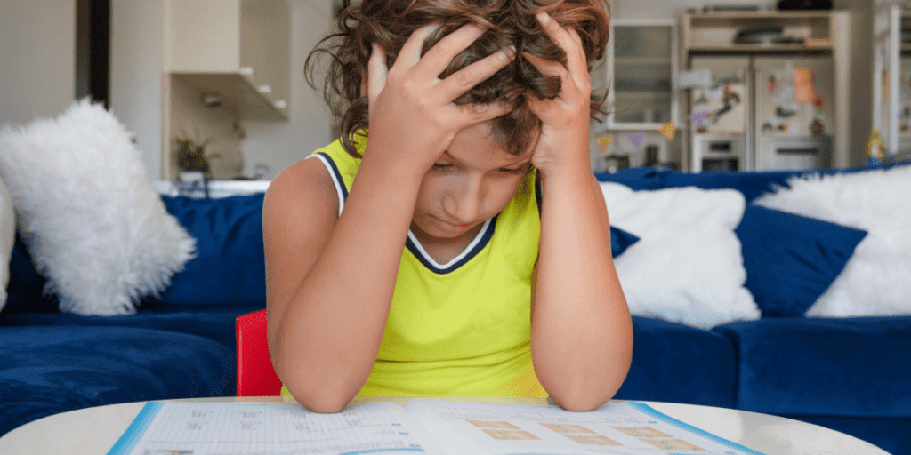 Child in yellow shirt looks frustrated with schoolwork at a white table indoors.
