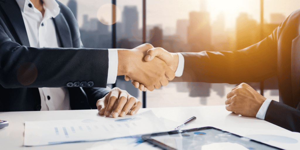 Two people in suits shaking hands over a Simplified table with documents and a tablet.