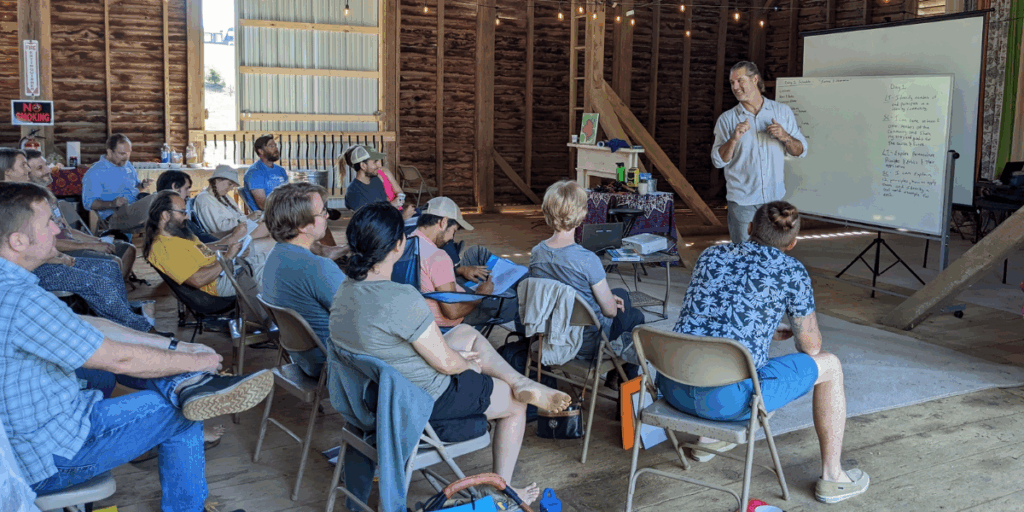 A group maps their inner landscape and build relationships as Ryan Blosser presents by a whiteboard in a rustic setting.