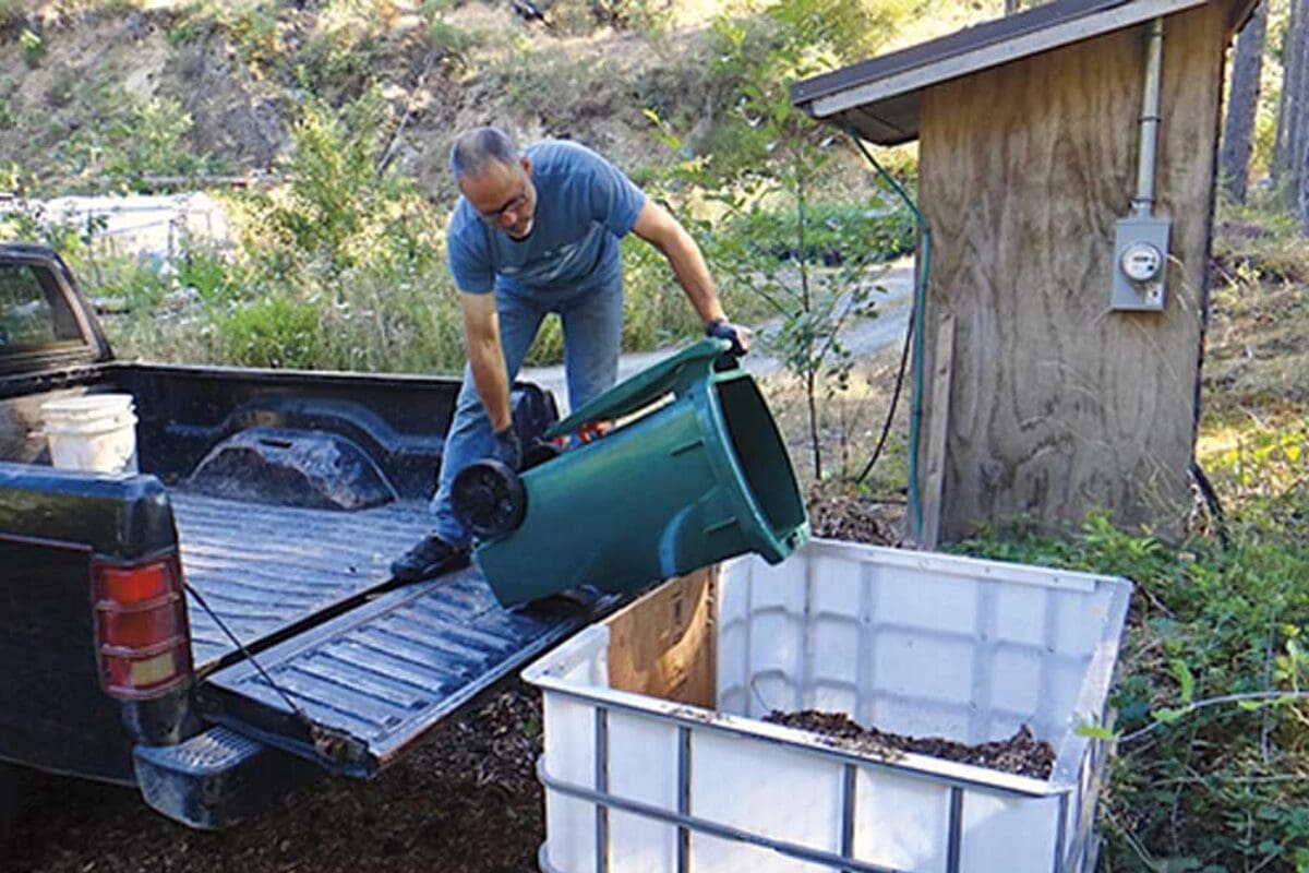 Man empties green bin of organic waste into large white container beside composting toilets.